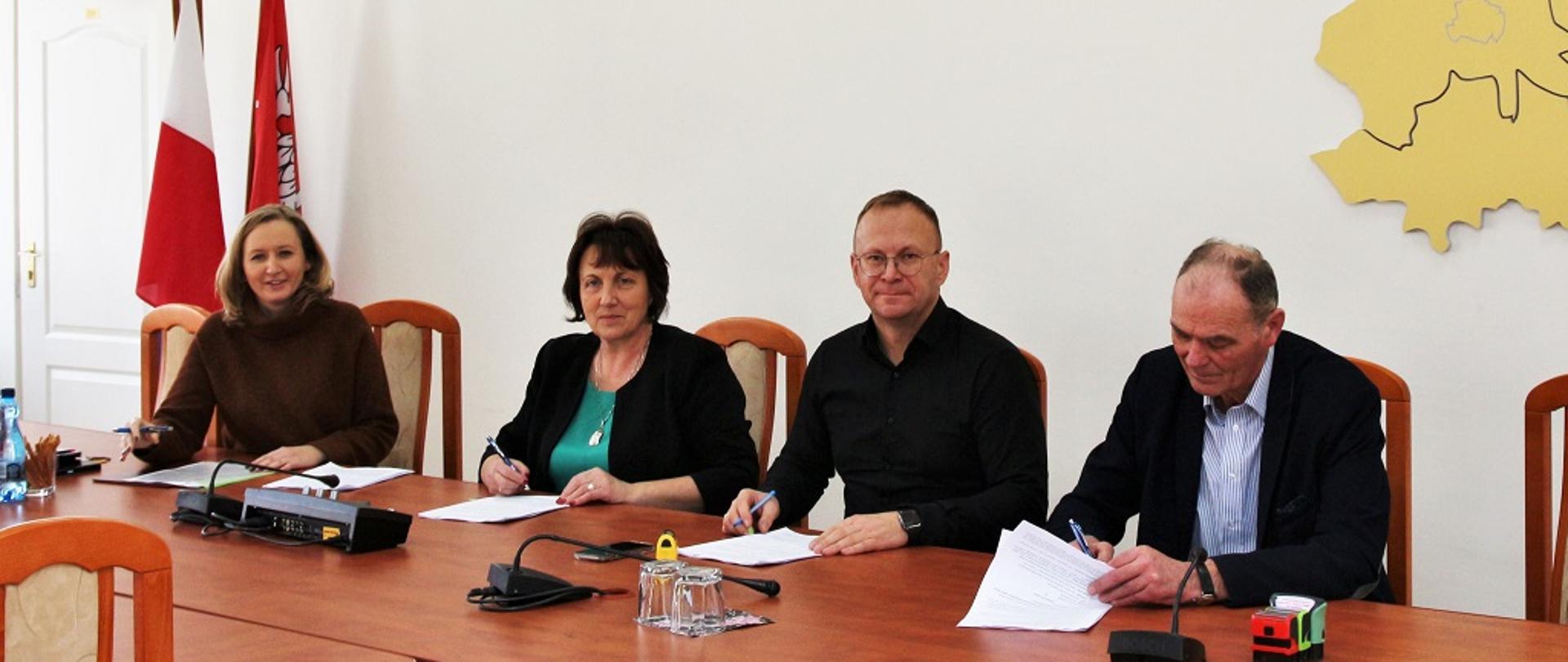 Na fotografii siedzących przy podłużnym stole od lewej: skarbnik powiatu Iwona Odrobińska, starosta Elżbieta Sadowska, właściciel firmy P.W. Wikrusz, wicestarosta Ryszard Domański. Jest to moment składania podpisów na umowie. Nad podpisującymi na ścianie w tle złożony z przestrzennych liter napis Powiat Sokołowski wraz z mapą obrazującą obszar powiatu, krzyż, oraz złoty orzeł w koronie. W tle flagi polski oraz województwa mazowieckiego.