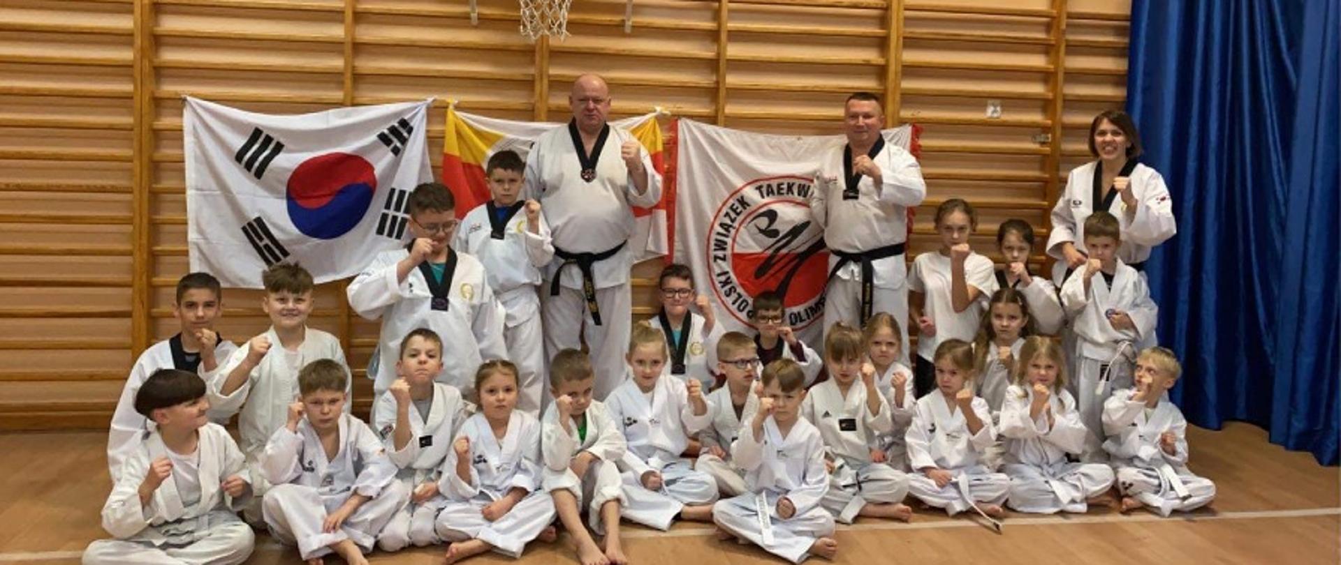 Dzieci w białych kimonach siedzą w rzędzie na podłodze. Za nimi stoją dorośli. Na ścianie z drabinek rozwieszone są flagi