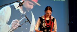 kobieta w bogato zdobionym gorsecie, w białej bluzce i czerwonych koralach na szyi gra na cymbałach 