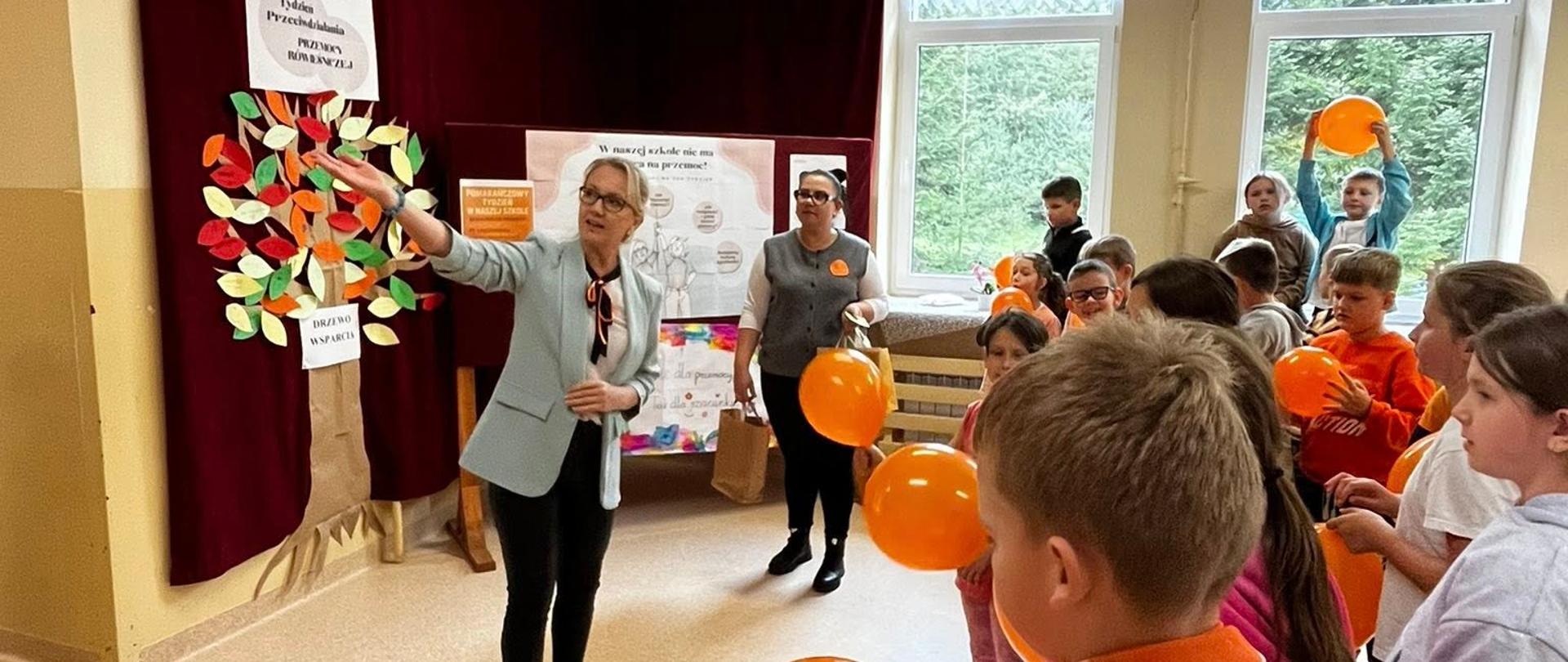 Na środku korytarza szkolnego pani dyrektor szkoły wskazuje placem hasła dotyczące przeciwdziałania przemocy, po prawej stronie stoi pani psycholog, trzyma w ręku pomarańczowy balon, na ścianie na bordowej zasłonie zawieszone zostało wycięte z papieru jesienne drzewo z kolorowymi liśćmi, przed panią dyrektor stoją uczniowie, trzymają w rękach pomarańczowe balony