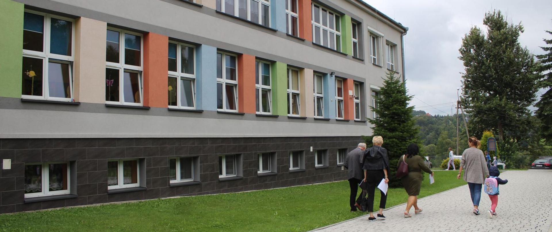 Na zdjęciu budynek szkoły w Zebrzydowicach, widoczna kolorowa elewacja i grupa osób w tle. 