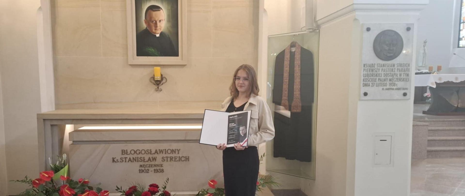 Kinga stoi przed grobem błogosławionego księdza Stanisława Streicha, trzyma w ręku dyplom laureatki, przed grobem znajdują się czerwone kwiaty, nad grobem wisi portret błogosławionego, obok w szklanej gablocie zawieszona szata księdza