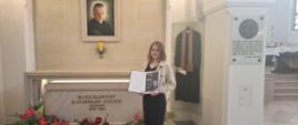 Kinga stoi przed grobem błogosławionego księdza Stanisława Streicha, trzyma w ręku dyplom laureatki, przed grobem znajdują się czerwone kwiaty, nad grobem wisi portret błogosławionego, obok w szklanej gablocie zawieszona szata księdza
