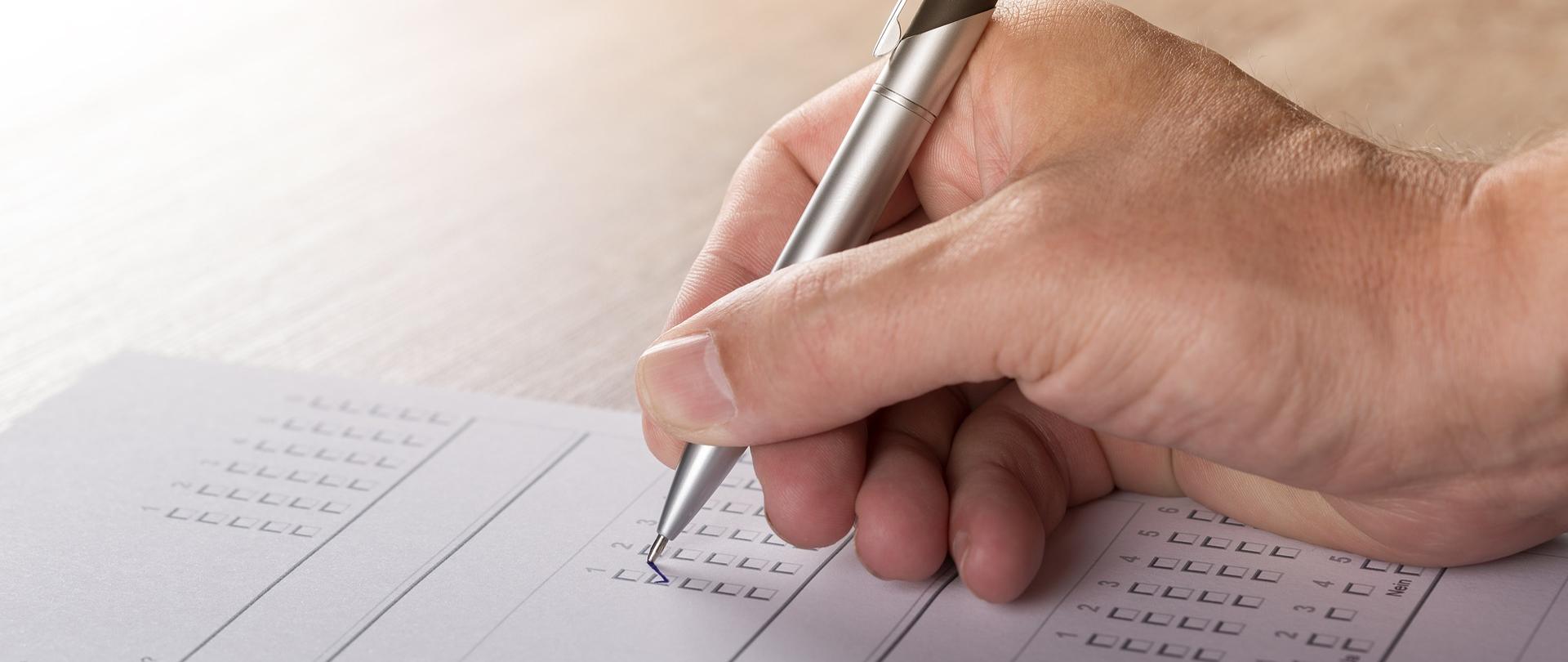 Zdjęcie przedstawia kartkę papieru, a na niej dłoń trzymającą długopis, zaznaczającą odpowiednią kratkę na kartce.