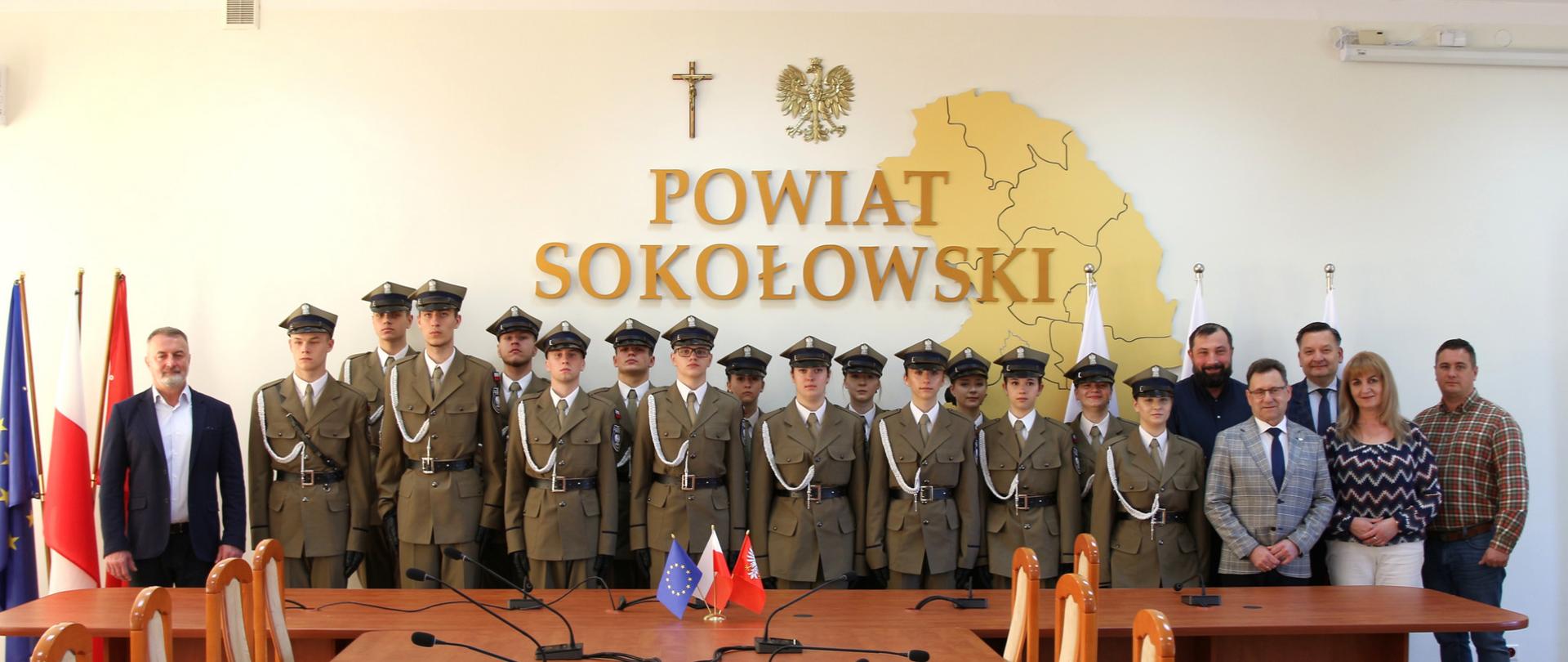 Zdjęcie grupowe zwycięzców wraz z członkami Zarządu Powiatu na tle ściany z przestrzennym napisem Powiat Sokołowski. Ba ścianie widzimy także krzyż katolicki, orła z godła i przestrzenną mapę z zarysem granic powiatu.