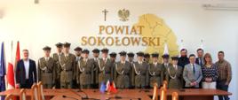 Zdjęcie grupowe zwycięzców wraz z członkami Zarządu Powiatu na tle ściany z przestrzennym napisem Powiat Sokołowski. Ba ścianie widzimy także krzyż katolicki, orła z godła i przestrzenną mapę z zarysem granic powiatu.