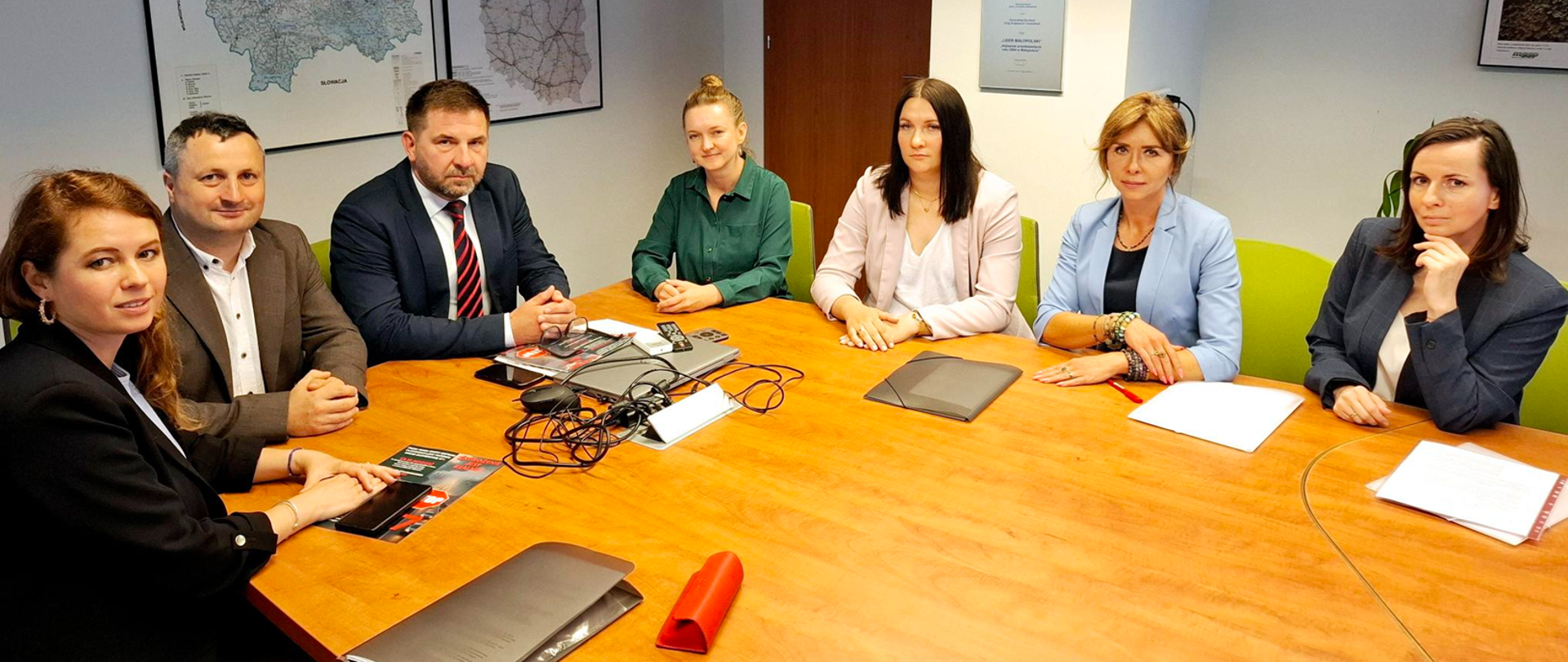 Siedem osób – pięć kobiet i dwóch mężczyzn – siedzi przy stole konferencyjnym w sali biurowej. W tle widoczne są mapy i certyfikaty na ścianach. Na stole leżą dokumenty, laptopy i przewody. Wszyscy uczestnicy patrzą w stronę obiektywu. Atmosfera spotkania jest formalna.