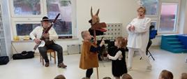 Zdjęcie prezentuje dzieci biorące udział w audycji muzycznej oraz teatrzyku. Przed grupą siedzących dzieci występują aktorzy