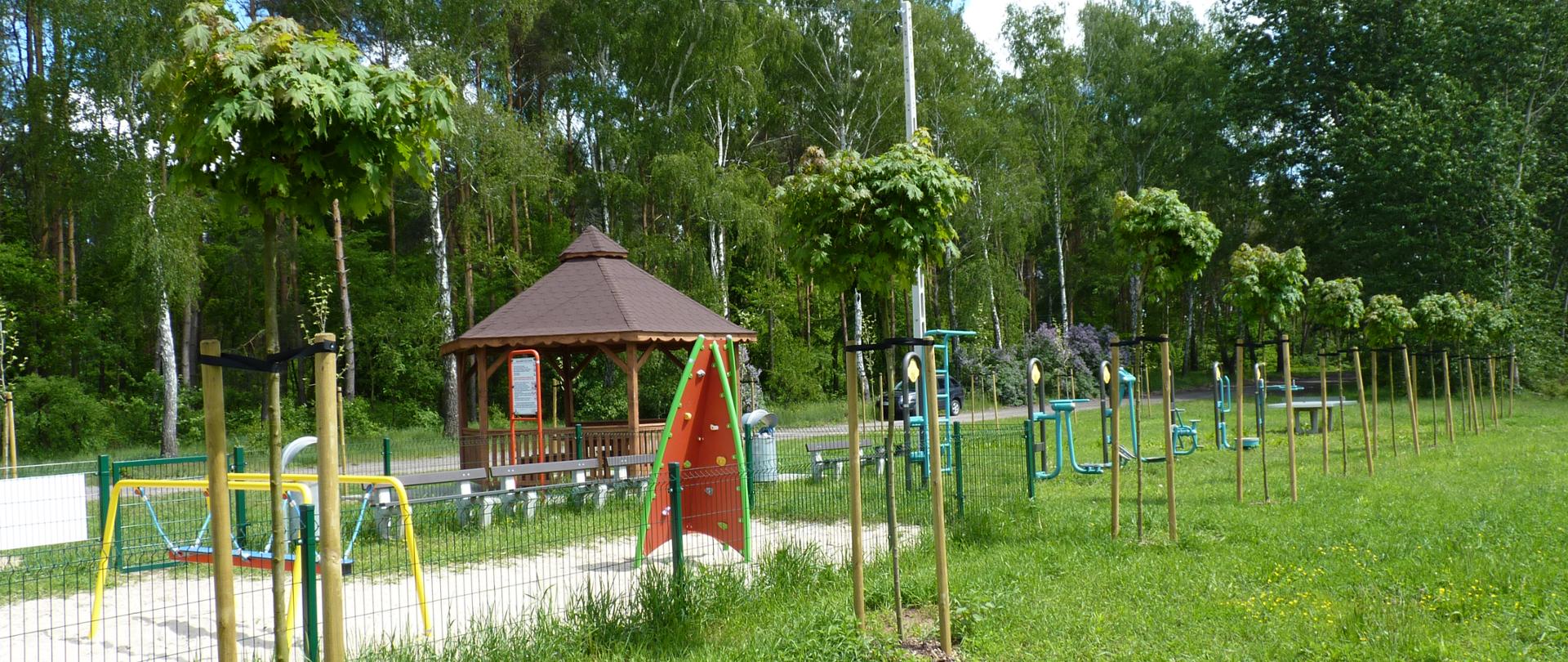 Młode drzewka z palikami podtrzymującymi po dwóch stronach. W tle plac zabaw i brązowa altana