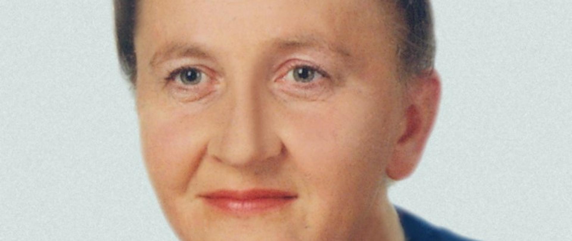 Starsza kobieta, na tym zdjęciu pokazana jako szatynka. Ma jasne oczy, długie włosy opięte w kok. Ubrana w granatowy sweter z widocznym dekoltem w serek, a pod spodem koronkowa biała bluzka