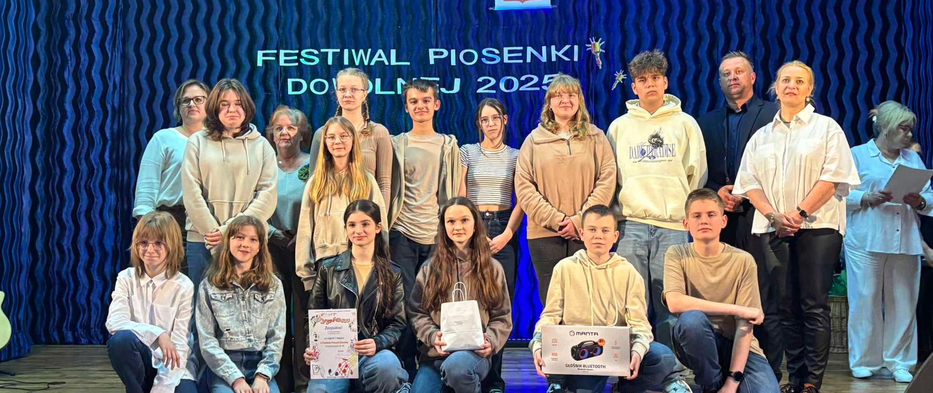 Grupa uczniów, którzy reprezentowali naszą szkołę w konkursie pozuje do wspólnego zdjęcia. W dłoniach trzymają dyplomy i nagrody (torebkę prezentową oraz karton z radiem). Z tyłu znajduje się granatowe tło, a na nim napis: FESTIWAL PIOSENKI DOWOLNEJ 2025.