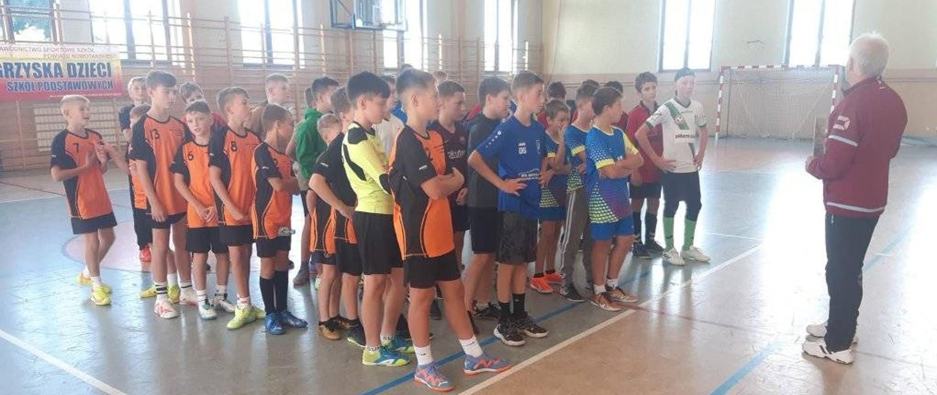 Uczniowie ustawieni w rzędach na hali sportowej, gotowi drużynowo do rozpoczęcia igrzysk.