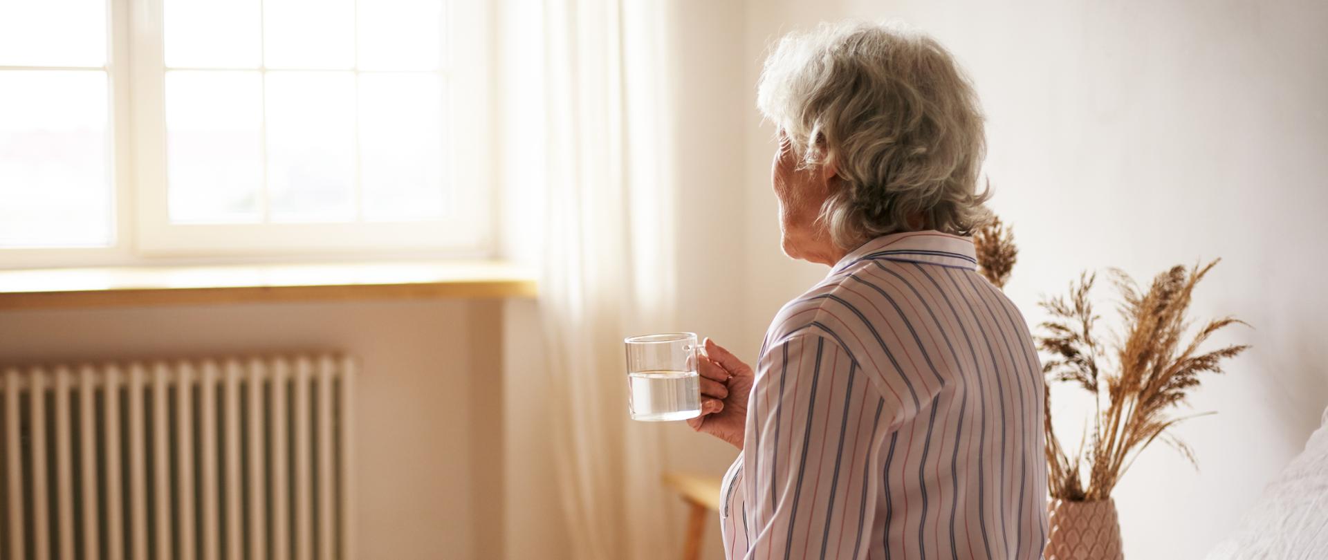 Rear view of senior sixty year old woman with gray hair holding mug washing down sleeping pill, suffering from insomnia. Elderly retired female taking medicine with water, sitting in bedroom