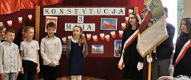 Dzieci występujące w akademii z okazji 232. rocznicy uchwalenia Konstytucji 3 Maja, biało czerwona flaga i dekoracje, sztandar szkoły, dzieci oglądające występ