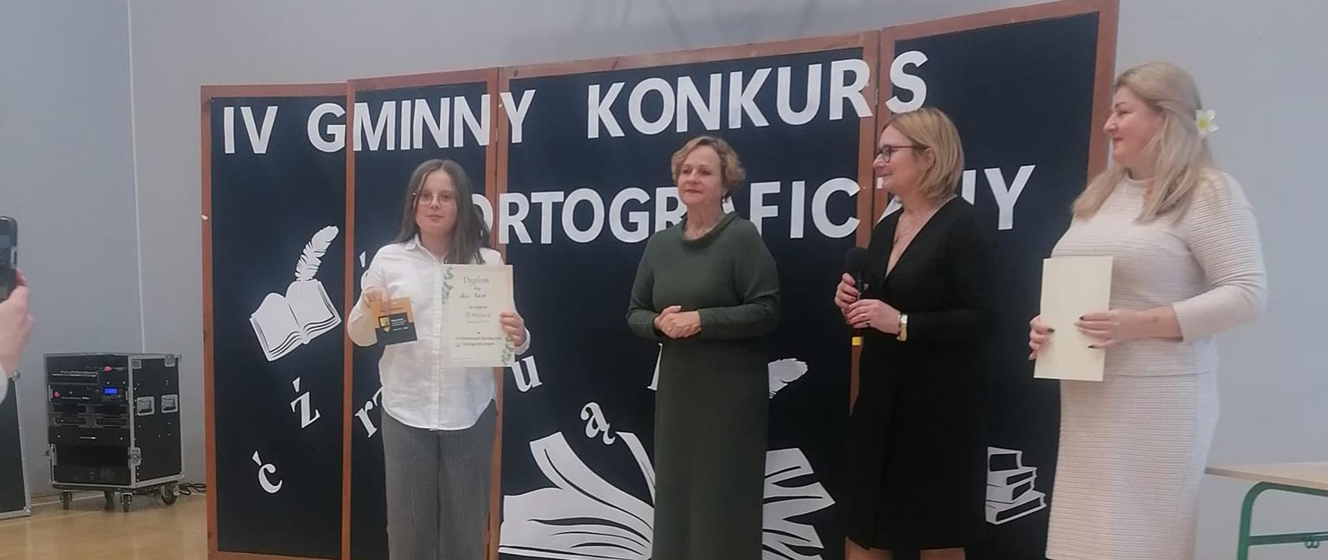 Maja w szarych spodniach, białej koszuli stoi z dyplom i ze statuetką stoi obok jury na sali gimnastycznej.