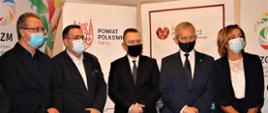 Czterech mężczyzn z lewej strony oraz jedna kobieta z prawej strony, stoją w maseczkach na tle kolorowych banerów.