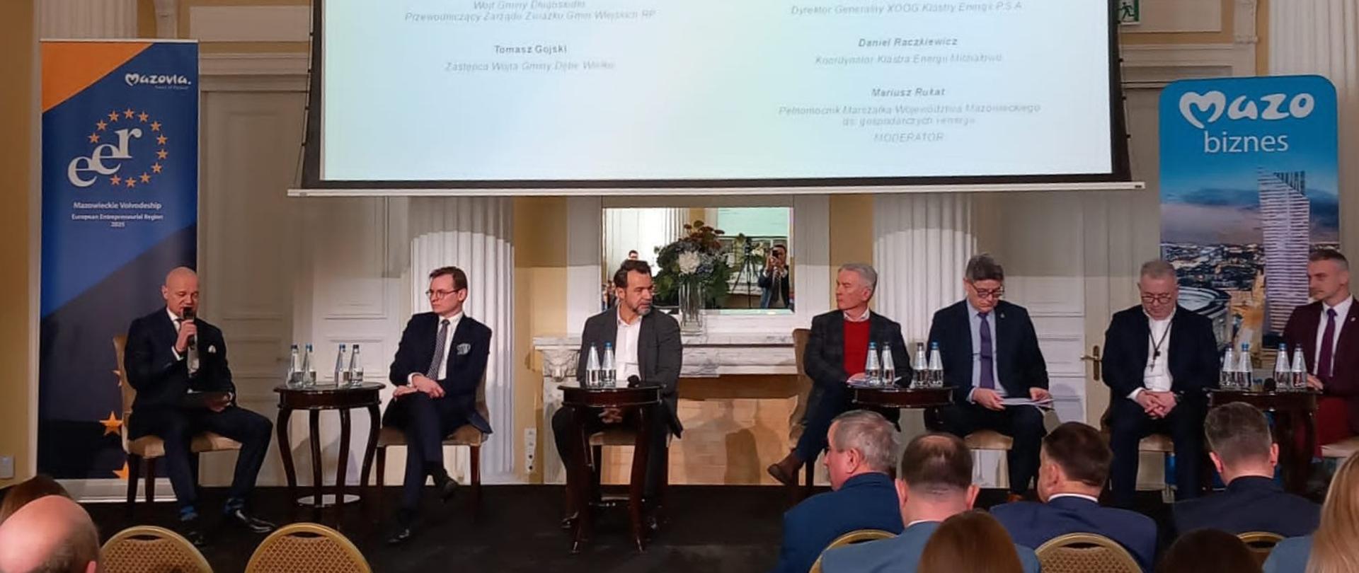 Na zdjęciu trwający panel dyskusyjny. Na scenie w eleganckiej sali z białymi kolumnami siedzi grupa osób prowadzących dyskusję. Nad nimi wisi ekran, na którym prezentowany jest tytuł dyskusji "Energetyka rozproszona w środowisku samorządów - wyzwania, możliwości, potrzeby". Przed sceną widownia składająca się z obecnych na Forum uczestników.