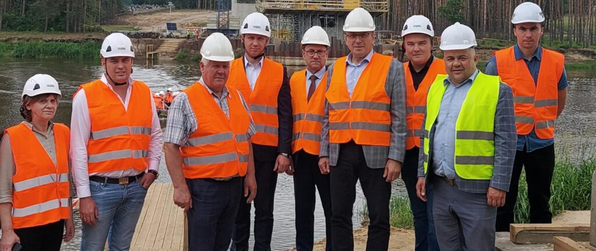 Zdjęcie grupowe uczestników spotkania na tle rzeki Bug. W tle betonowe elementy powstającego mostu.