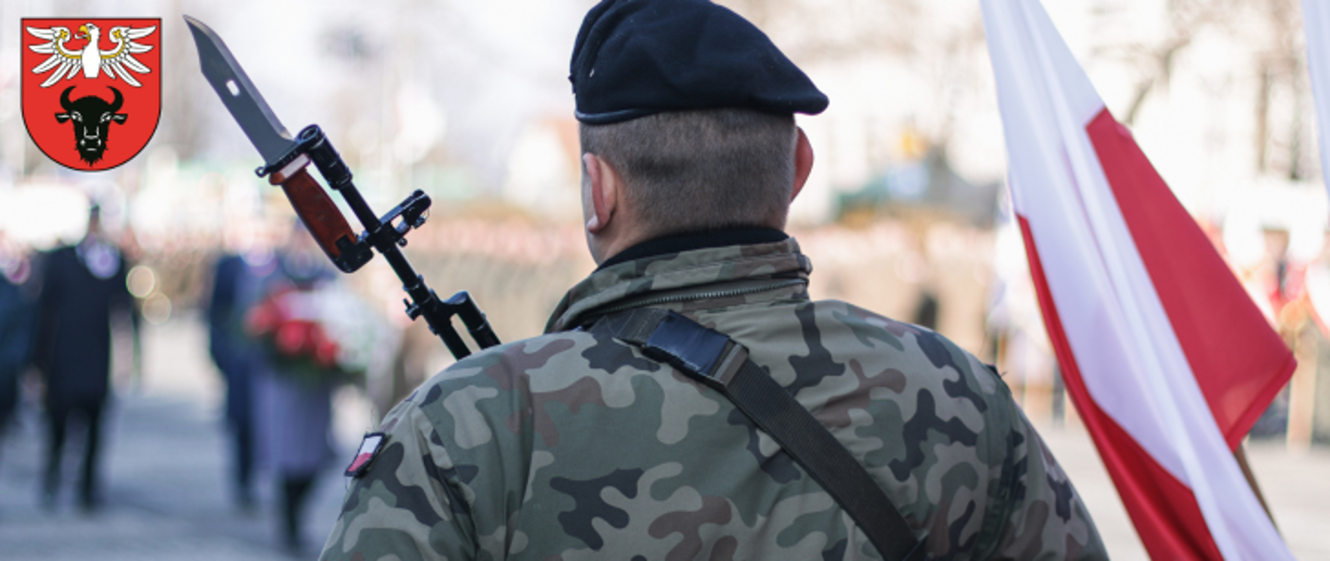 zdjęcie żołnierza trzymającego w ręku broń, w tle flaga biało-czerwona, w lewym górnym rogu herb powiatu