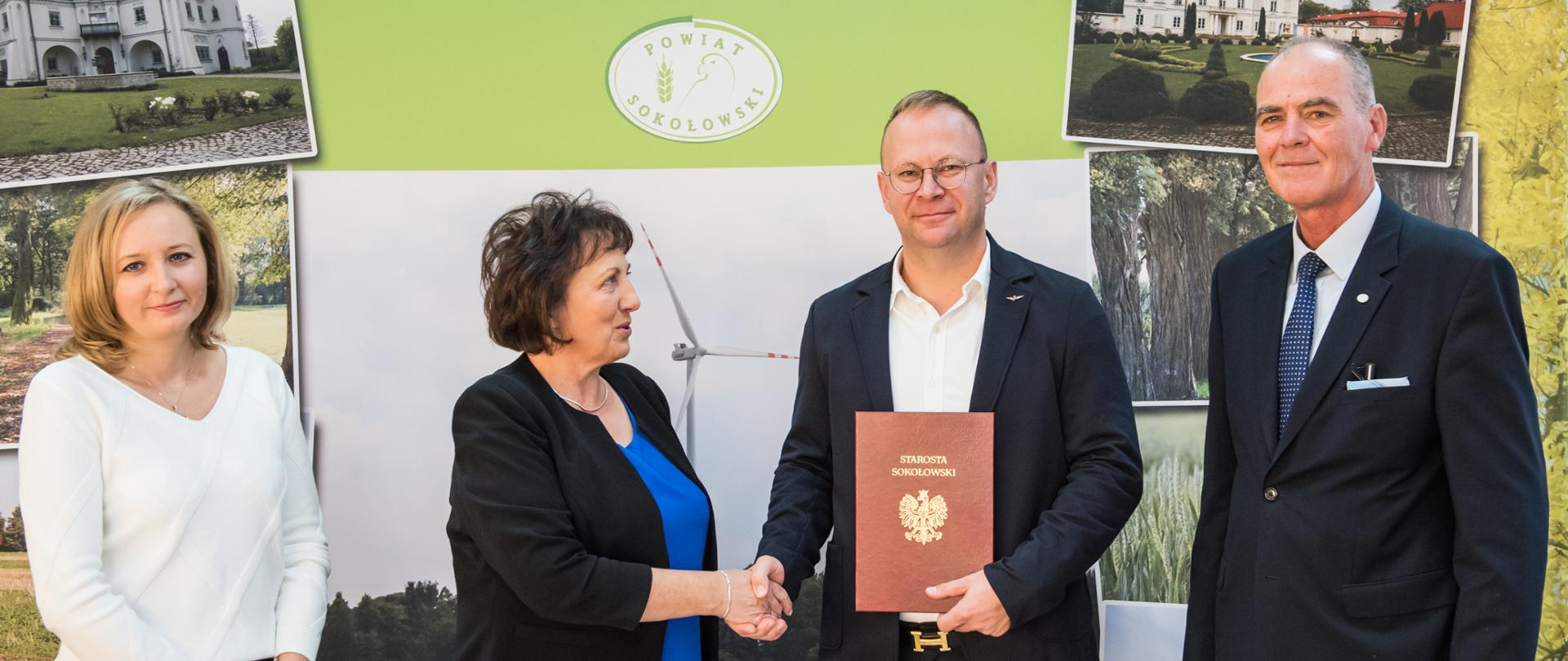 Zdjęcie grupowe. Na pierwszym planie od lewej skarbnik powiatu, starosta sokołowski, wykonawca zadania, wicestarosta. W tle ścianka z logo powiatu sokołowskiego oraz zdjęciami miejsc z terenu powiatu.