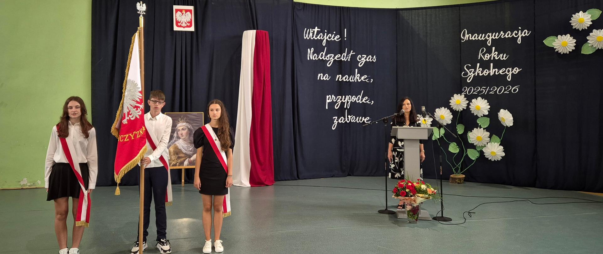 Chłopiec i dwie dziewczyny stoją w dużej sali na baczność. Są przepasani biało czerwonymi wstęgami. Chłopiec trzyma sztandar. Dalej przy szarej mównicy stoi kobieta. Za nią granatowa ścianka z napisem Inauguracja roku szkolnego i białymi kwiatami 