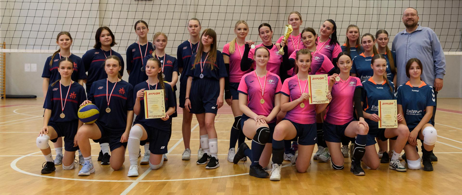 Fotografia grupowa na hali sportowej drużyn od lewej prezentuje się drużyna w granatowych koszulkach, następnie w różowych zwyciężczynie z pucharem oraz drużyna w ciemnoniebieskich koszulkach oraz przewodniczący Rady Powiatu Sokołowskiego.