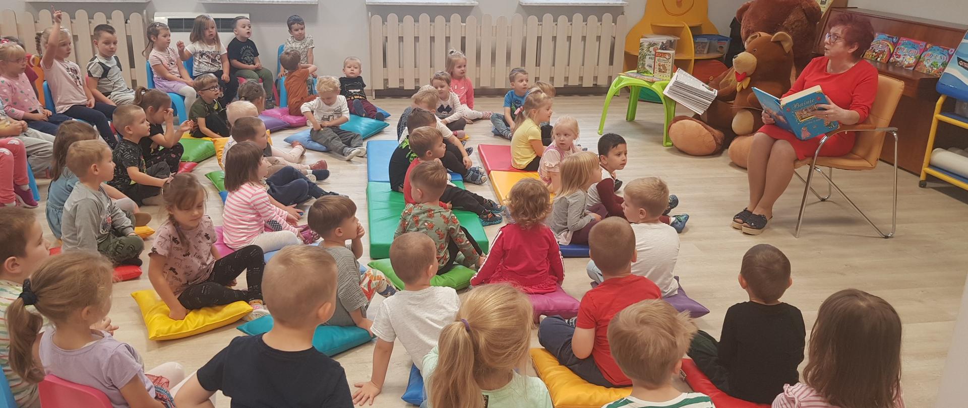 Siedząca grupa dzieci słucha opowiadania czytanego przez nauczyciela 