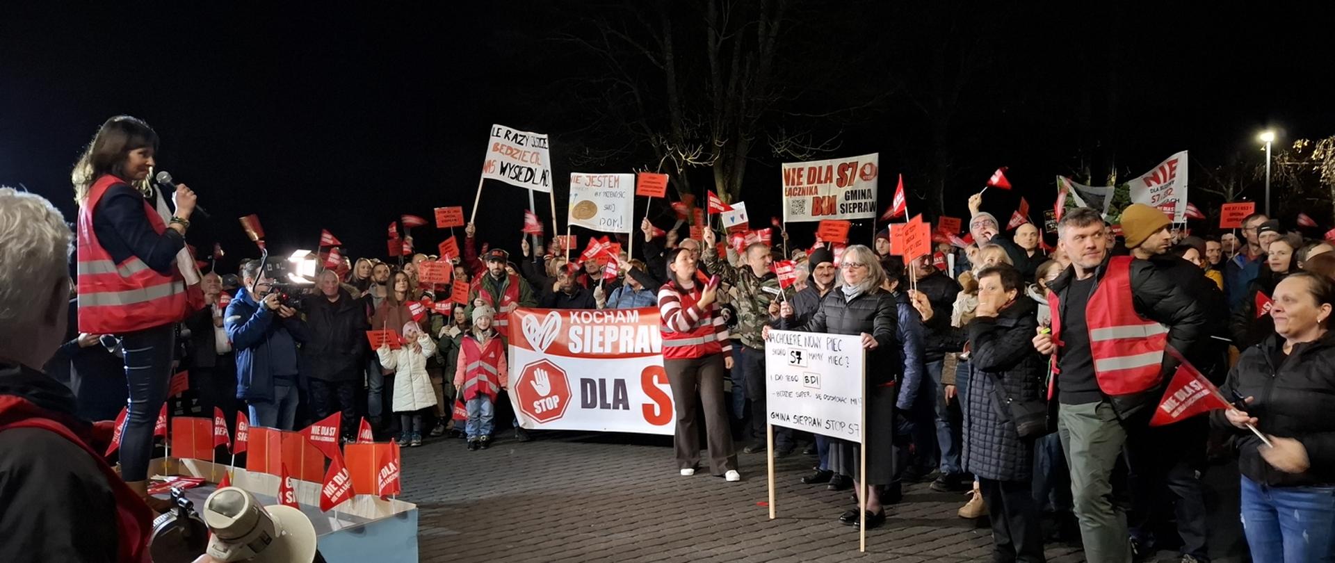 Podwyższone stanowisko z osobą przemawiającą jest otoczone liczną grupą mieszkańców z flagami i transparentami. W tle widoczne są drzewa i oświetlenie uliczne, a atmosfera jest dynamiczna i głośna.
