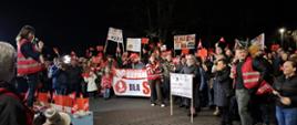 Podwyższone stanowisko z osobą przemawiającą jest otoczone liczną grupą mieszkańców z flagami i transparentami. W tle widoczne są drzewa i oświetlenie uliczne, a atmosfera jest dynamiczna i głośna.