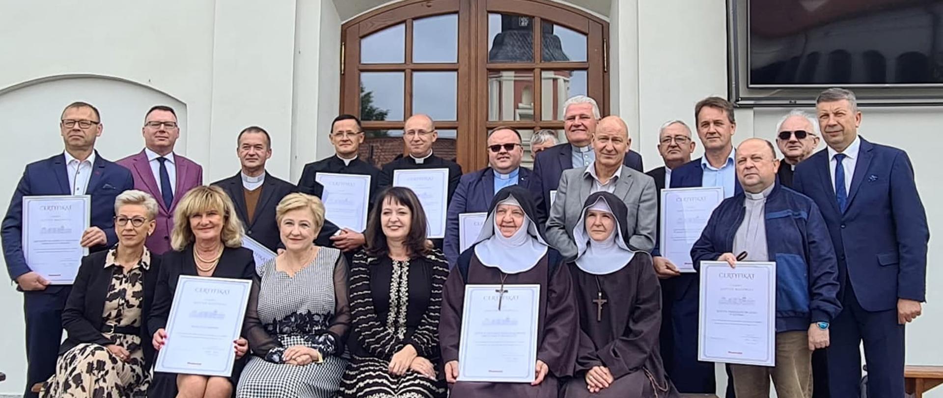 Zdjęcie wykonano w malowniczej scenerii na dziedzińcu przed Sanktuarium Św. Antoniego Padewskiego w Ostrołęce. W pierwszym rzędzie, w centrum zasiadają Przełożona Klasztoru Mniszek Klarysek Kapucynek, Siostra Daria. Na prawo od Siostry Darii siedzi Siostra Klara, na lewo Członkinie Zarządu Województwa Mazowieckiego – Janina Ewa Orzełowska i Elżbieta Lanc. Za Siostrą Klarą stoi Dyrektor Wydziału Budownictwa w przasnyskim Starostwie - Jarosław Włodarczyk, a skrajnie z prawej strony Radny Województwa Mazowieckiego Mirosław Augustyniak. Dookoła pozostali uczestnicy, którzy podobnie jak Klaryski Kapucynki otrzymali certyfikat "Cenny Zabytek Mazowsza".