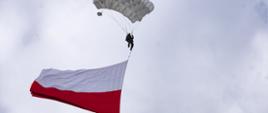 spadochroniarz na tle chmur skaczący ze spadochronem, mający przywiązaną do siebie rozwiniętą biało czerwoną flagę