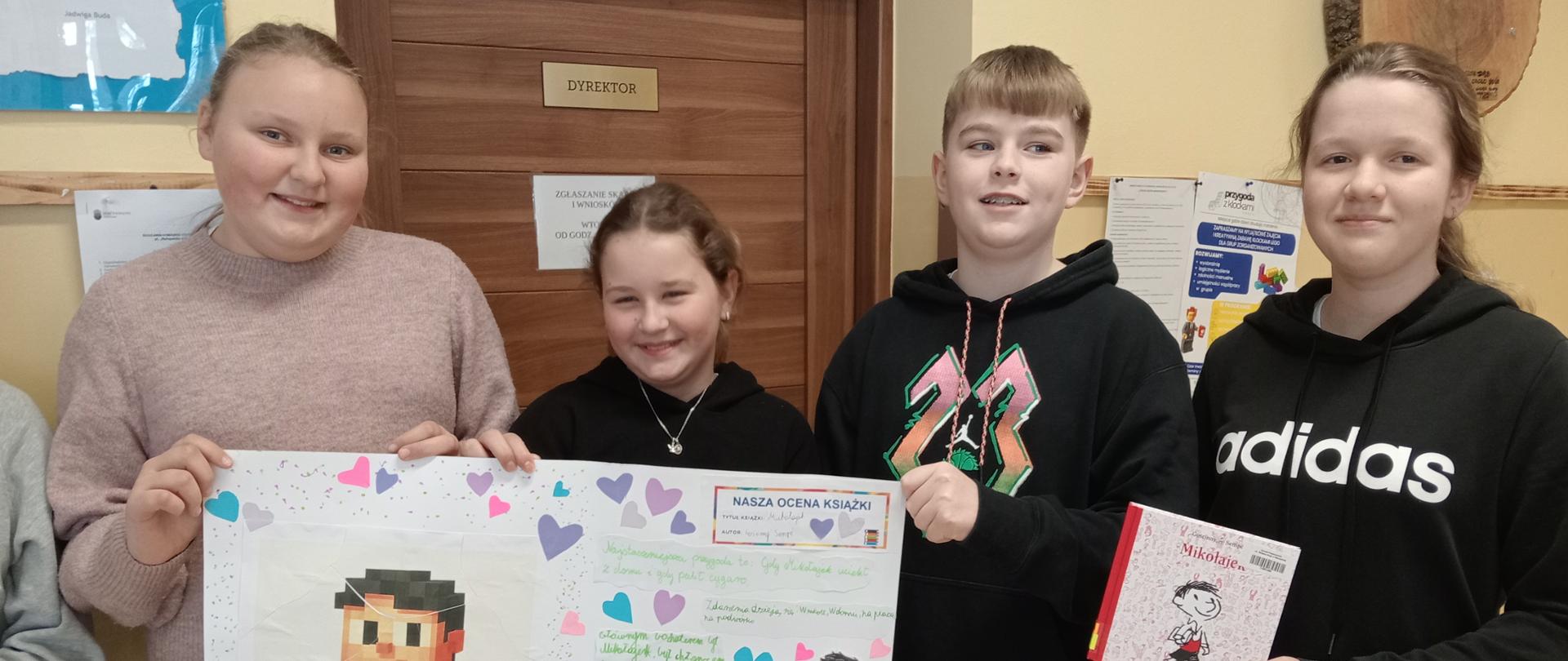 Trzy dziewczynki i chłopiec stoją pod ścianą na korytarzu szkolnym i trzymają wspólnie wykonany grupowo duży plakat z postacią Mikołajka, bohatera lektury "Mikołajek", dziewczynka stojąca po lewej trzyma w ręku książkę "Mikołajek"
