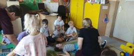 Na dywanie leży dwoje dzieci - chłopiec i dziewczynka. Nad chłopcem pochyla się uczeń klasy ósmej, nad dziewczynką uczennica klasy ósmej. Wokół nich, na dywanie siedzą przedszkolaki. Twarze dzieci są skierowane na uczniów klasy ósmej.
