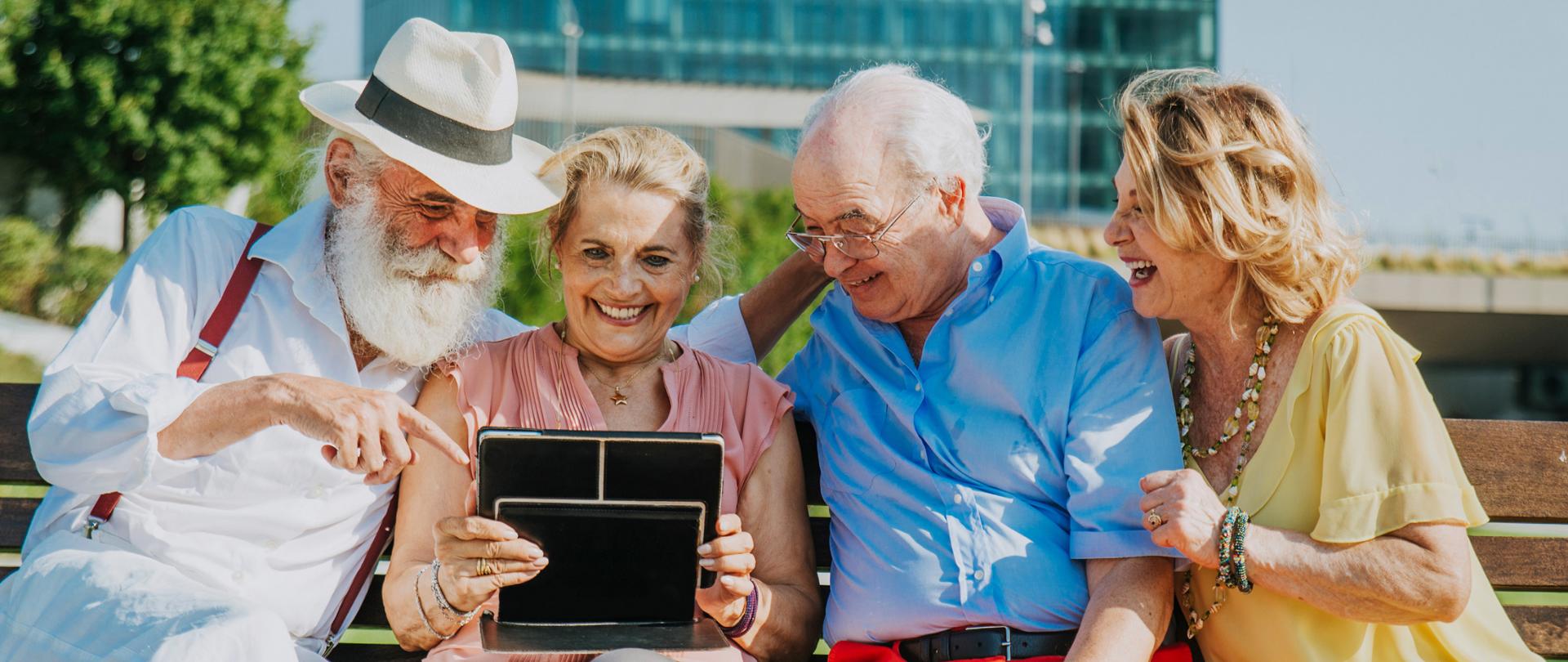 Uśmiechnięta czwórka seniorów siedzi na ławce w mieście i patrzy na tablet trzymany przez jedną z osób