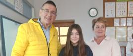 Na zdjęciu znajduje się troje osób. W środku stoi uczennica klasy szóstej, która trzyma w dłoniach zieloną torebkę prezentową. Po jej prawej stronie znajduje się mężczyzna, a po lewej - Pani Dyrektor. Wszyscy z uśmiechami pozują do wspólnego zdjęcia.