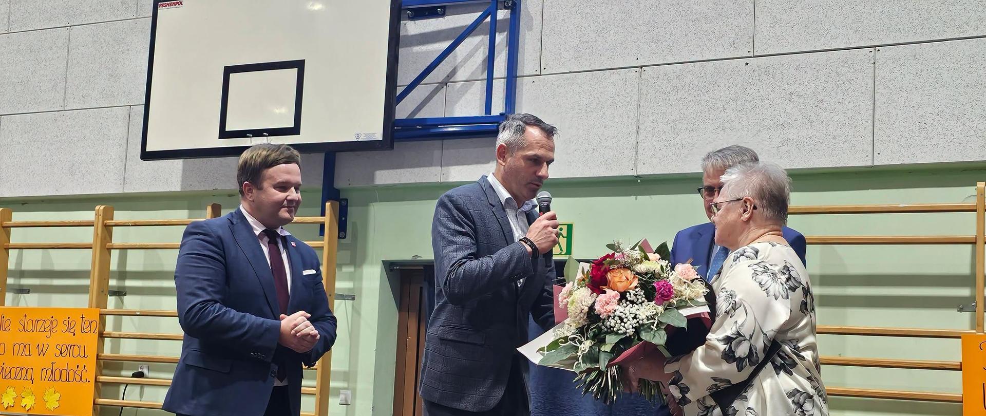 Grupa osób stojąca na scenie; jedna z nich wręcza bukiet kwiatów innej osobie, a dwie pozostałe przyglądają się tej scenie.