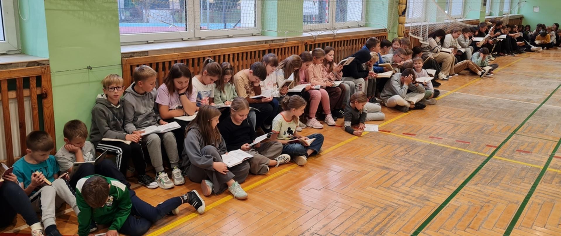 Grupa dzieci siedzi na sali gimnastycznej i każdy czyta swoją ulubioną książkę.