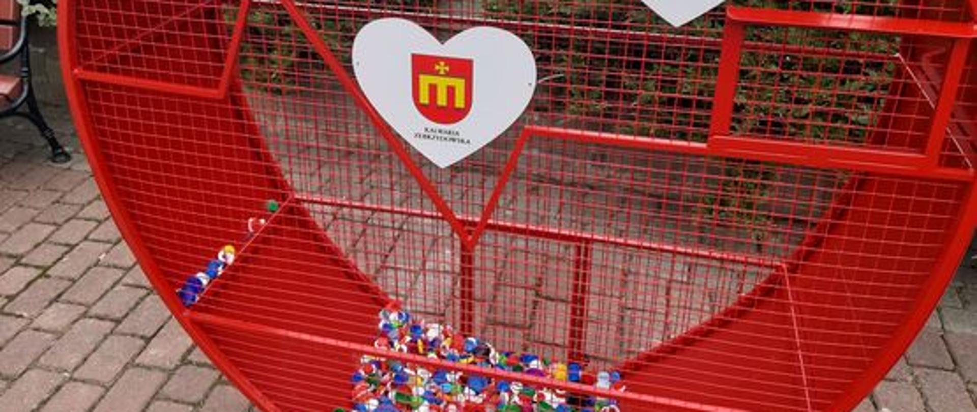 Metalowy czerwony pojemnik z siatki w kształcie serca na kostce brukowej. Wewnątrz widać plastikowe nakrętki. W tle żywopłot oraz parking z samochodami. 