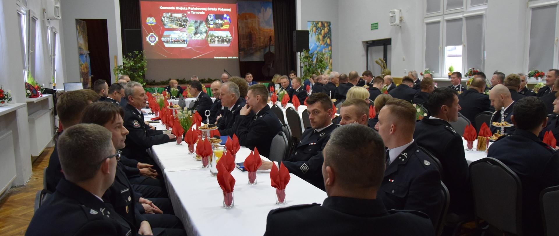 Sala wypełniona umundurowanymi strażakami OSP i PSP siedzącymi przy długich stołach podczas oficjalnej odprawy operacyjnej w Wietrzychowicach.