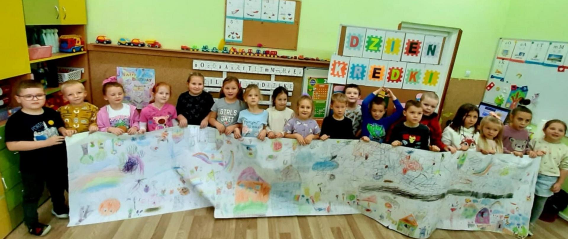 Grupka dzieci pozuje do zdjęcia stojąc w szeregu i trzymając w dłoniach kilkumetrowy zwój papieru, na którym widnieją ich rysunki. W tle napis Dzień Kredki.