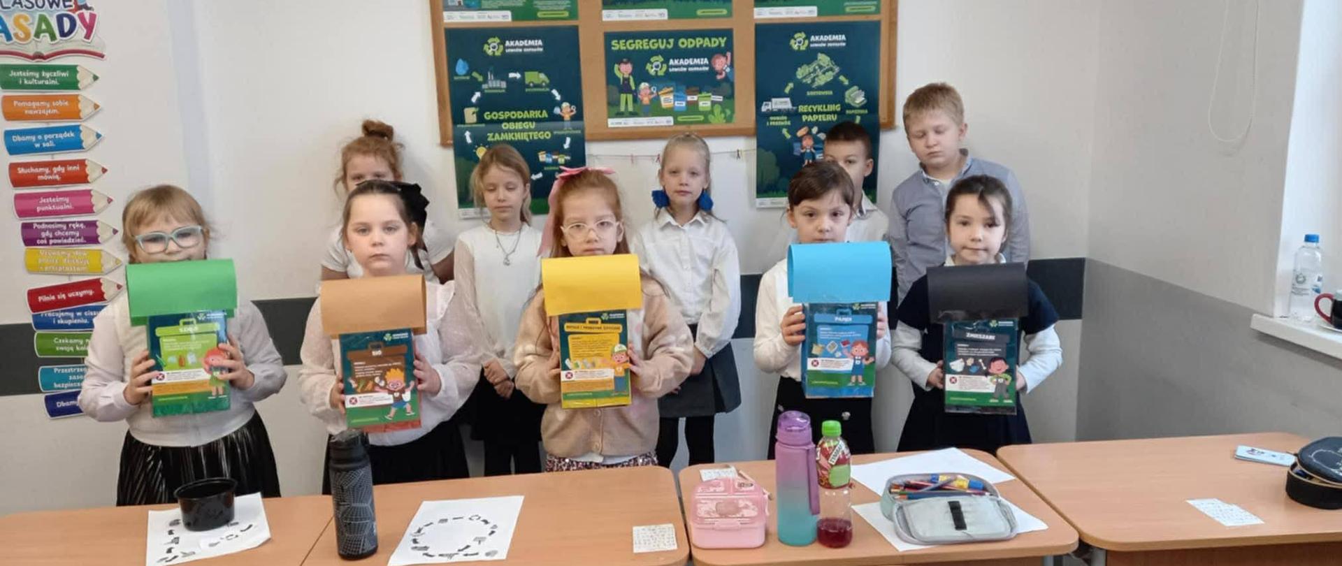 Dziesięcioro uczniów klasy pierwszej pozuje do wspólnego zdjęcia. Dzieci znajdują się w sali lekcyjnej. Uczniowie stoją w dwuszeregu. Dzieci stojące w pierwszym szeregu trzymają w dłoniach kartoniki w pięciu różnych kolorach: zielonym, brązowym, żółtym, niebieskim i czarnym.