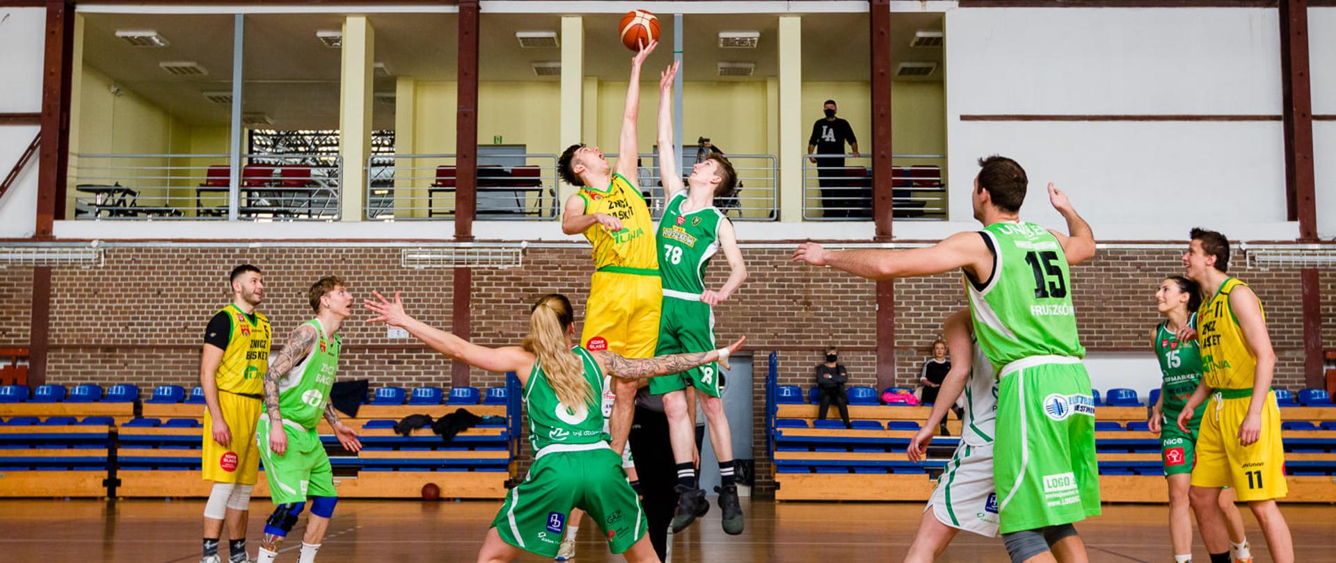 Na zdjęciu dwie drużyny koszykarzy. Drużyna w żółtych i drużyna w zielonych strojach rozgrywają mecz koszykówki. 