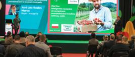 „Wykład na konferencji poświęconej uprawie truskawek. Prelegenci stoją na scenie przed dużym ekranem prezentacyjnym, na którym wyświetlane są informacje o narzędziu do zarządzania pozostałościami środków ochrony roślin. Publiczność siedzi na krzesłach i słucha wystąpienia.”