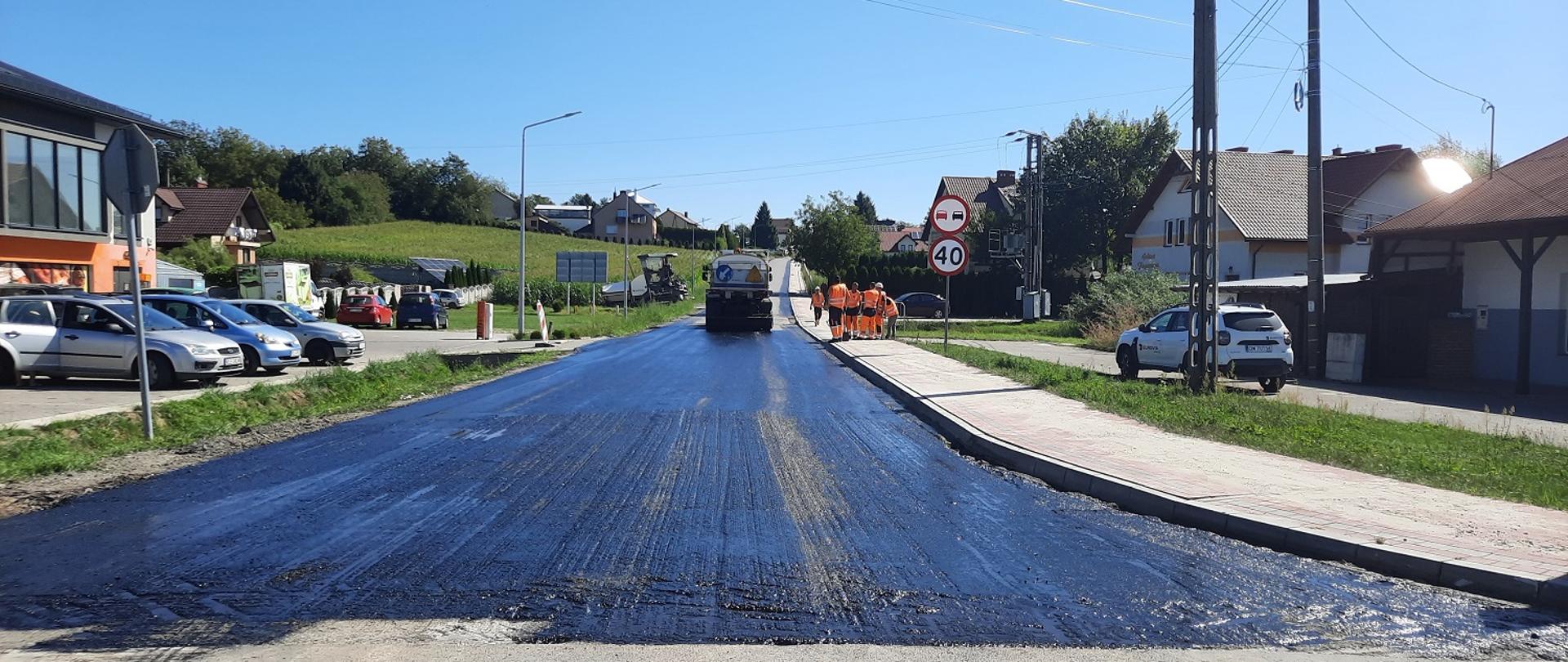 remontowana droga, czarna warstwa ścieralna wylewana na drogę na całej szerokości w tle robotnicy w pomarańczowych strojach i duże auto ciężarowe 