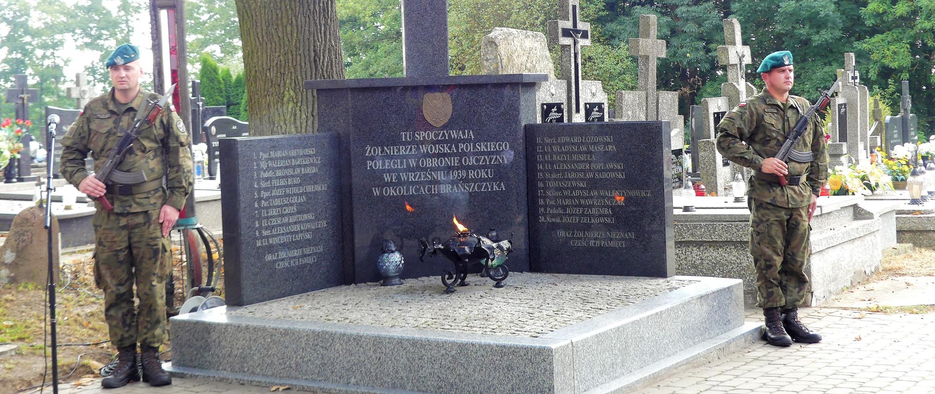 Dwóch żołnierzy stojących obok zbiorowej mogiły żołnierzy poległych w obronie ojczyzny we Wrześniu 1939 roku w okolicach Brańszczyka. W tle inne mogiły zmarłych osób.
