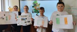 Czwórka uczniów stoi w rzędzie w klasie szkolnej, prezentując swoje wykonane odręcznie plakaty o Irlandii. Od lewej stoją dwie dziewczynki, a po nich dwóch chłopców. Pierwsze dwa plakaty zawierają bogate opisy, zdjęcia i rysunki konturu Irlandii, natomiast dwaj chłopcy trzymają białe kartki z narysowaną flagą Irlandii (pionowe pasy: zielony, biały, pomarańczowy). W tle za dziećmi znajduje się duży interaktywny ekran, na którym wyświetlona jest grafika przedstawiająca uśmiechniętego leprechauna w zielonym kapeluszu i stroju. 