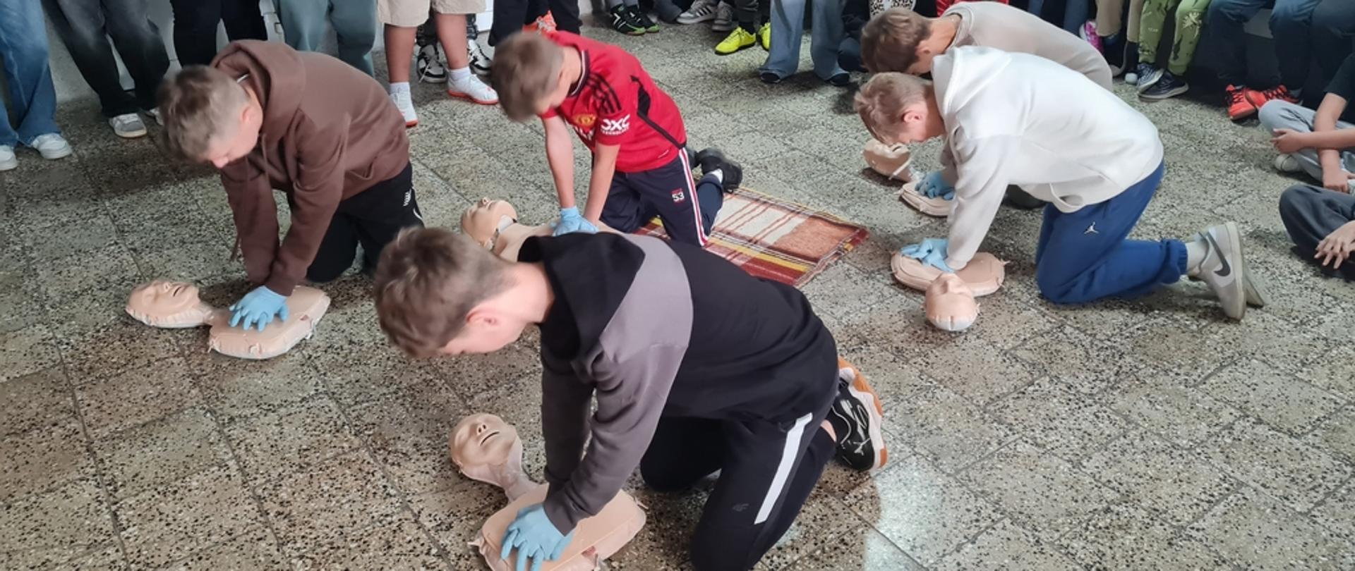 Na szkolnym korytarzu grupka dzieci w pozycji klęczącej ćwiczy resuscytację na leżących na ziemi fantomach. Inne dzieci stoją lub siedzą wokół i przyglądają się.