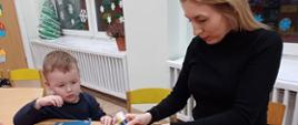 Chłopczyk i jego mama siedzą przy stole, wspólnie tworząc świąteczne ozdoby - papierową torbę z bożonarodzeniowym akcentem