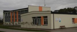 Zdjęcie przedstawia Szkołę Podstawową w Nowym Niestępowie. Budynek ma nowoczesną architekturę i jasną elewację z pomarańczowymi oraz szarymi elementami. Składa się z dwóch części – niższej z przodu i wyższej w tle. Na ścianach znajdują się duże okna, które wpuszczają dużo światła do środka. Przed budynkiem widoczny jest chodnik oraz ławka. W tle rosną drzewa, a nad budynkiem przebiegają linie energetyczne.