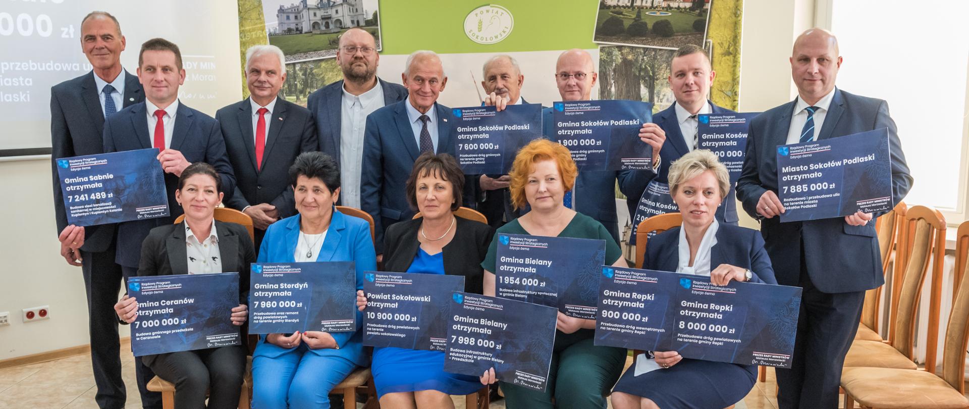 Zdjęcie grupowe na tle ścianki z logo Powiatu Sokołowskiego. Na zdjęciu wraz z parlamentarzystami wszyscy obecni na spotkaniu samorządowcy prezentujący swoje promesy, które zostały im wręczone. W pierwszym rzędzie na krzesłach siedzą panie, a z tyłu na stojąco panowie.
